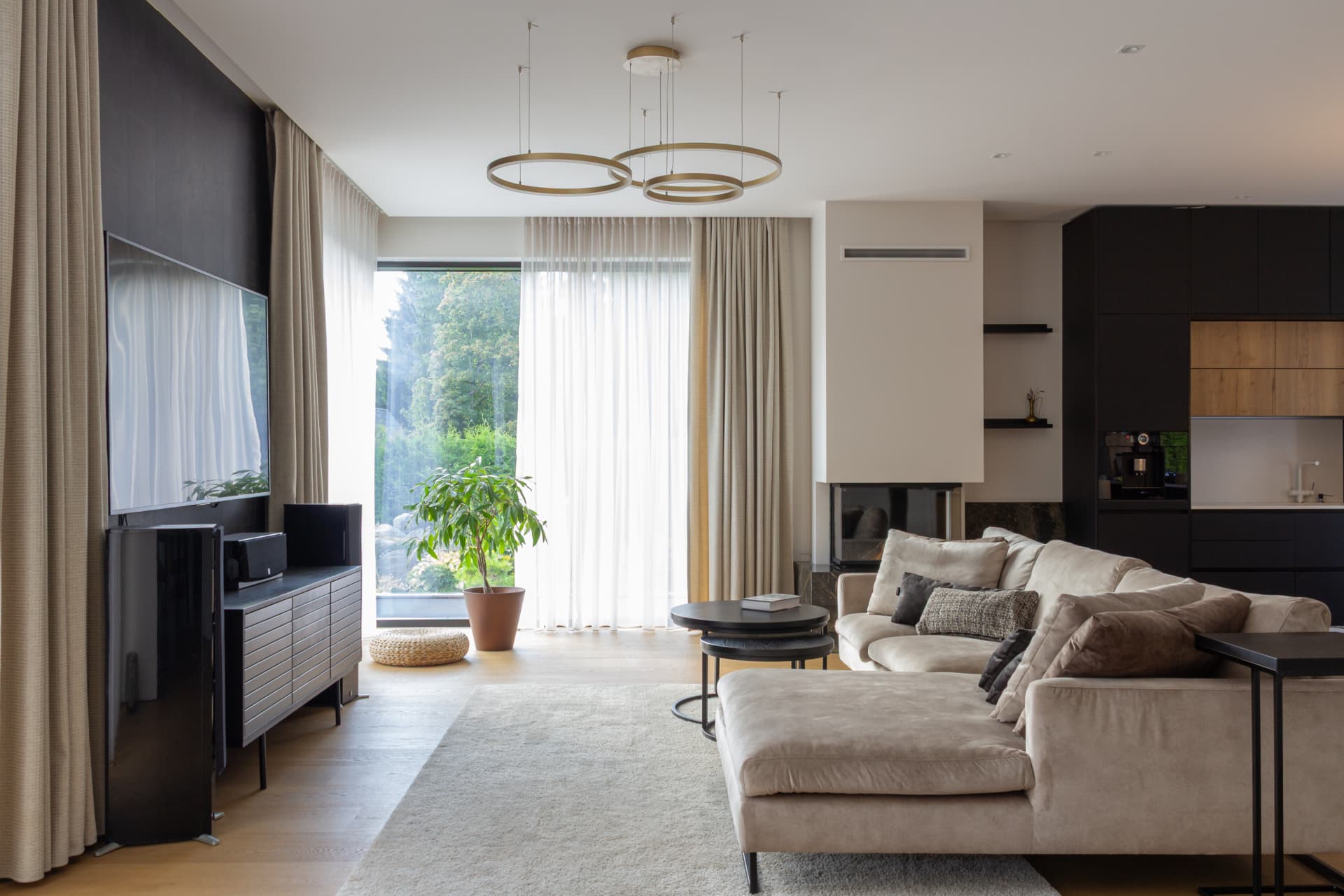 Open living room with a beige sofa, dark media wall, kitchen beyond and full-height windows